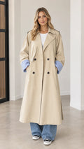 Front full-length view of woman wearing beige double-breasted maxi trench coat with blue contrast cuffs
