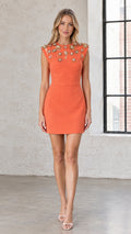 Front full view of woman wearing orange sleeveless crystal embellished mini sheath dress