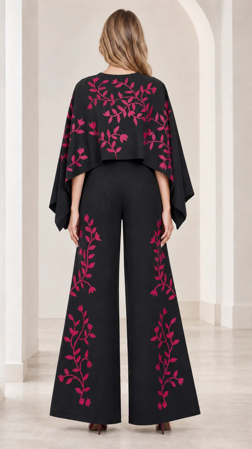Back view of model wearing black cape wide leg jumpsuit with pink vine embroidery